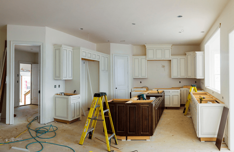 Kitchen under renovation with dusty subfloor, ladders, power tools, and construction debris